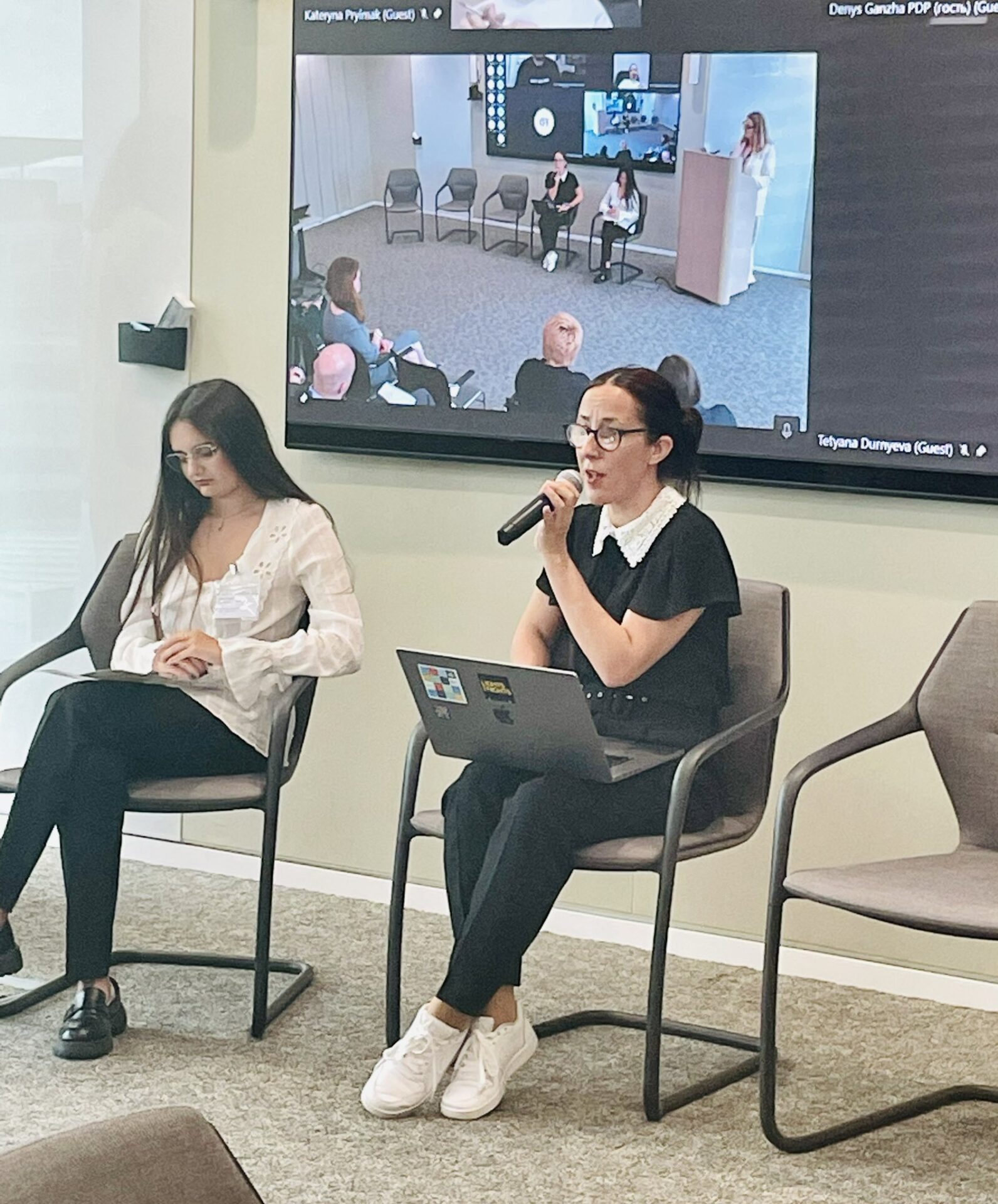 Юлія Сачук під час виступу на КонференціЇ з питань відновлення України 2023. Вона у чорні блузі та брюках, і окулярах. У неї зібране волосся. Вона сидить на стільці з ноутбуком на руках перед аудиторією слухачів_ок.