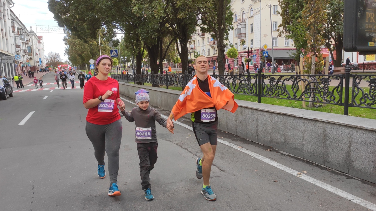 Дмитрик Ус бере участь у забігові в Чернігові разом з мамою і татом. Він біжить посередині, тримає за руки батьків. Мама в червоній футболці та сірих лосинах ліворуч, тато праворуч із помаранчевим прапором на плечах і сірих тренувальних шортах.