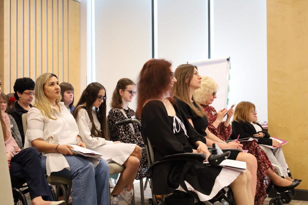 На фото дівчата та жінки сидять у профіль у два ряди, дивляться прямо. Скраю у першому ряду молода темноволоса кучеряа жінка на електричному кріслі колісному, це співачка Наталія Мартинова. На фоні 