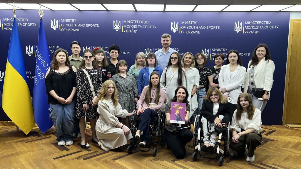 На фото близько 20 жінок і дівчат, двоє з них попереду на кріслах колісних, обабіч них навприсядки дві дівчини, а між ними жінка тримає в руках посібник з темнорожевою обкладинкою "Розвиток лідерства дівчат з інвалідністю". За жінками стоїть один високий русявий чоловік у блакитній сорочці. Це Владислав Яцук, представник Мінмолодьспорту. На фоні темно-синій банер із надписами - Міністерство молоді та спорту України (українською та англійською мовами). Ліворуч на фото два прапори - України та Мінмолодьспорту.