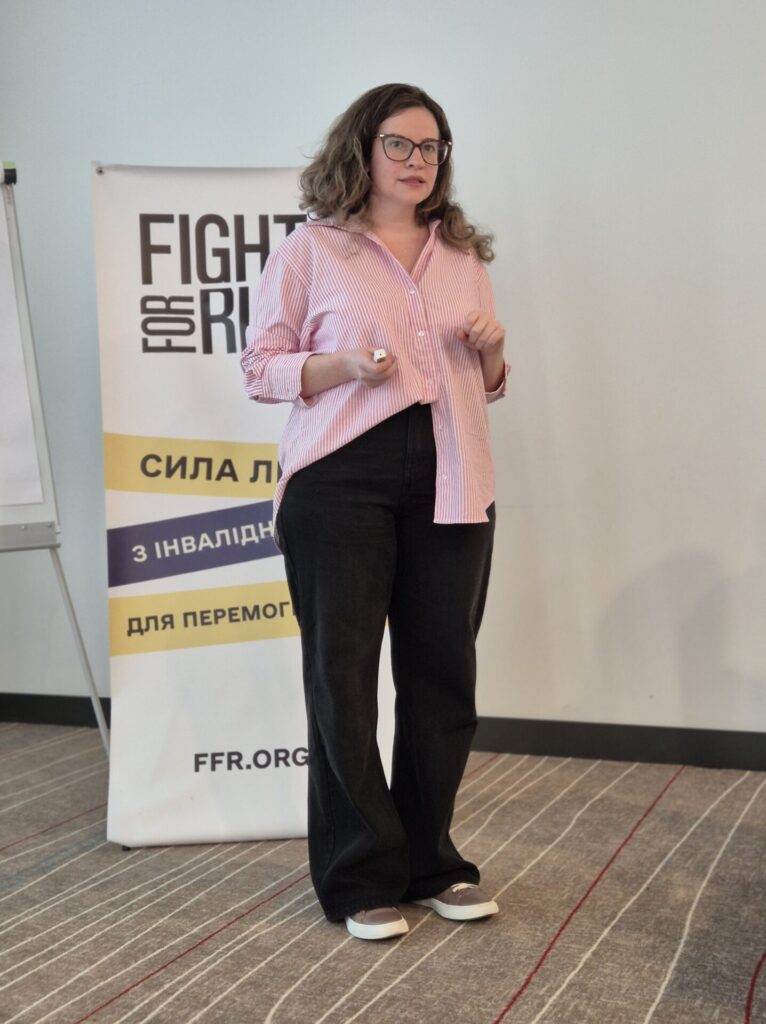 На фоні банера Fight For Right у повний зріст темнорусява жінка в окулярах. На ній рожева сорочка, темні широкі брюки і коричневі замшеві кросівки на світлій платформі.