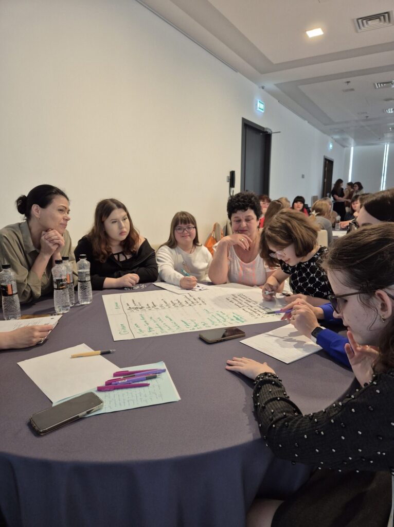 За круглим столом сидять дві жінки і четверо дівчат-тінейджерок. Одна з них пише щось на ватмані маркерами, а інші обговорюють, що саме потрібно писати. На передньому фоні столу лівіше лежать аркуші А4 із ручками та телефоном. 