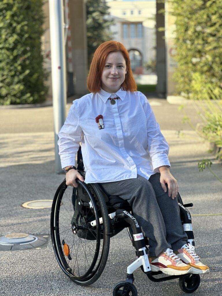 На кріслі колісному сидить молода жінка з волоссям до плечей красивого рудого кольору. На ній біла блуза з брошкою, сірі широкі брюки і світлі кросівки з жовтими смужками. Це Таня Герасимова. Фото зроблене на вулиці в сонячний день. 