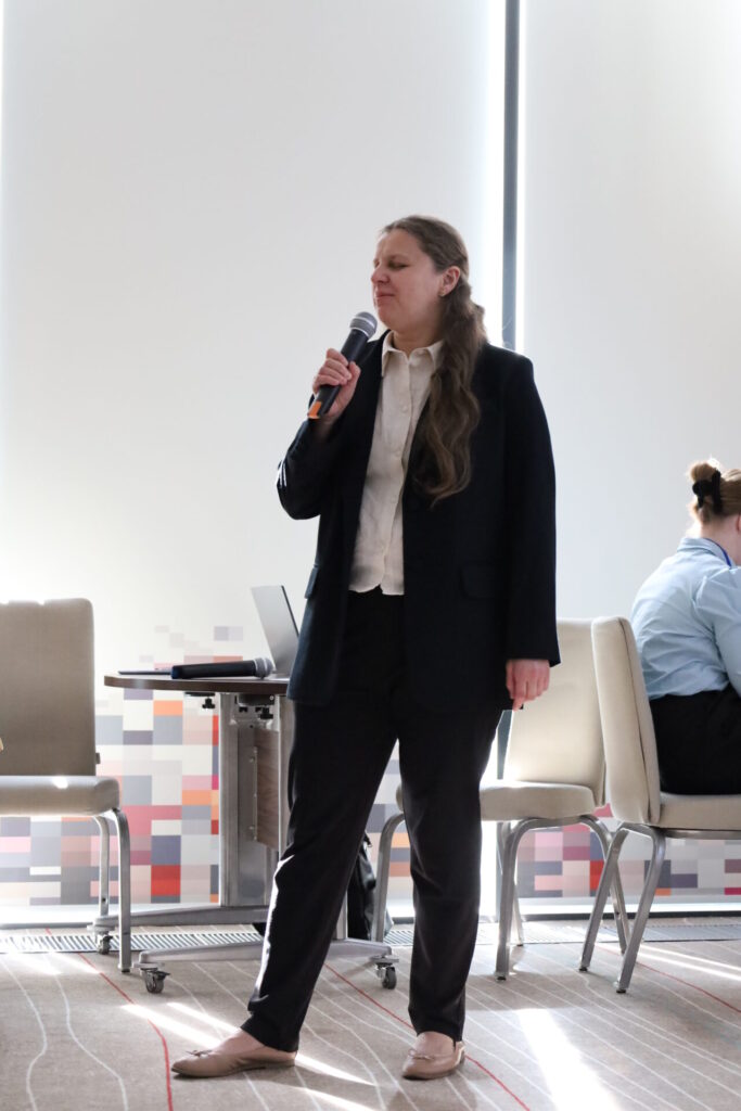 На фото жінка в повний зріст. У неї довге русяве волосся. Вона у темному брючному костюмі і світлій сорочці. Це Людмила Фурсова. Вона тримає мікрофон у правій руці і говорить.