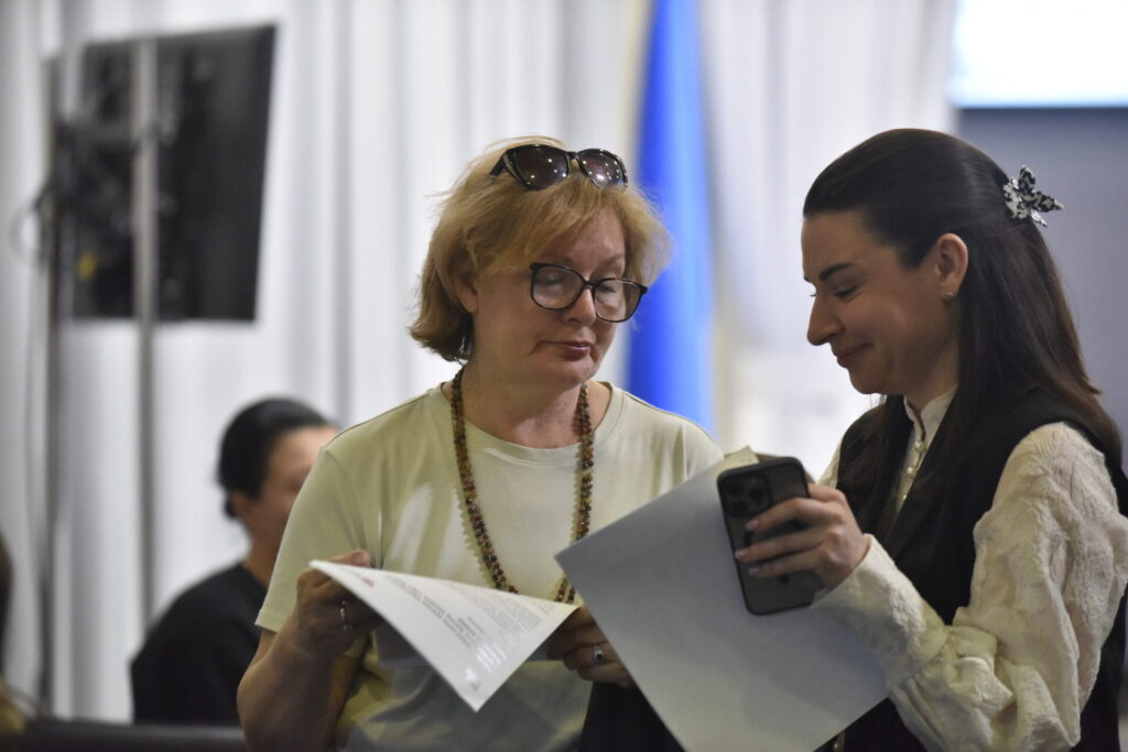 На фото праворуч темноволоса жінка у світлій блузі та жилеті. Це Марина Ткачук, представниця Національної ради з питань телебачення та радіомовлення спілкується зі світловолосою жінкою середнього віку. Вони обговорюють якісь документи, які тримають у руках.