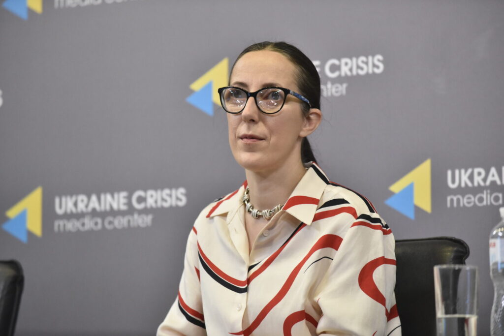 На зображенні Юлія Сачук сидить на фоні сірого банера Українського кризового медіацентру. Вона в окулярах у темній оправі, одягнена у світлу сорочку з червоними та чорними хвилястими смужками.