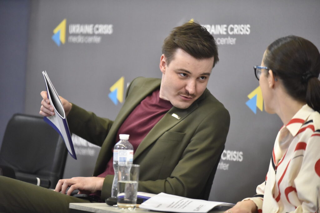 На зображенні темнорусявий чоловік середнього віку у темнозеленому костюмі повернувся до Юлії Сачук під час презентації і щось їй каже.