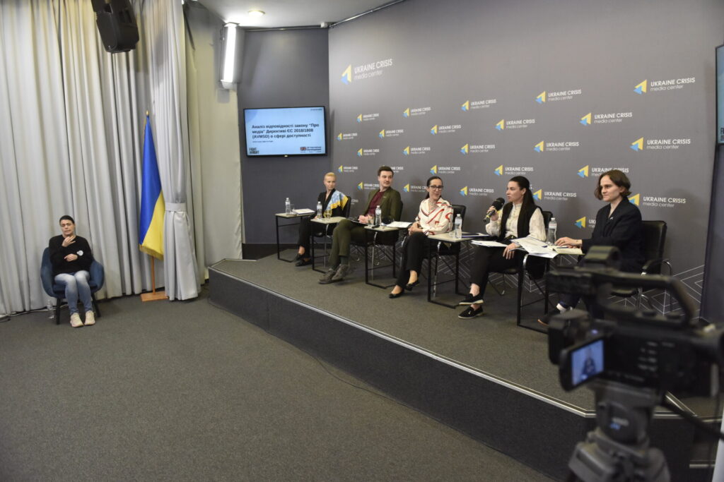На подіумі 4 спікерки та 1 спікер презентації та обговорення дослідження "Доступність аудіовізуальних медіапослуг в Україні: відповідність Закону "Про медіа" положенням Директиви ЄС 2018/1808". Прямо на фото сидить на стільці перекладачка жестової мови, біля неї стоїть прапоор України.