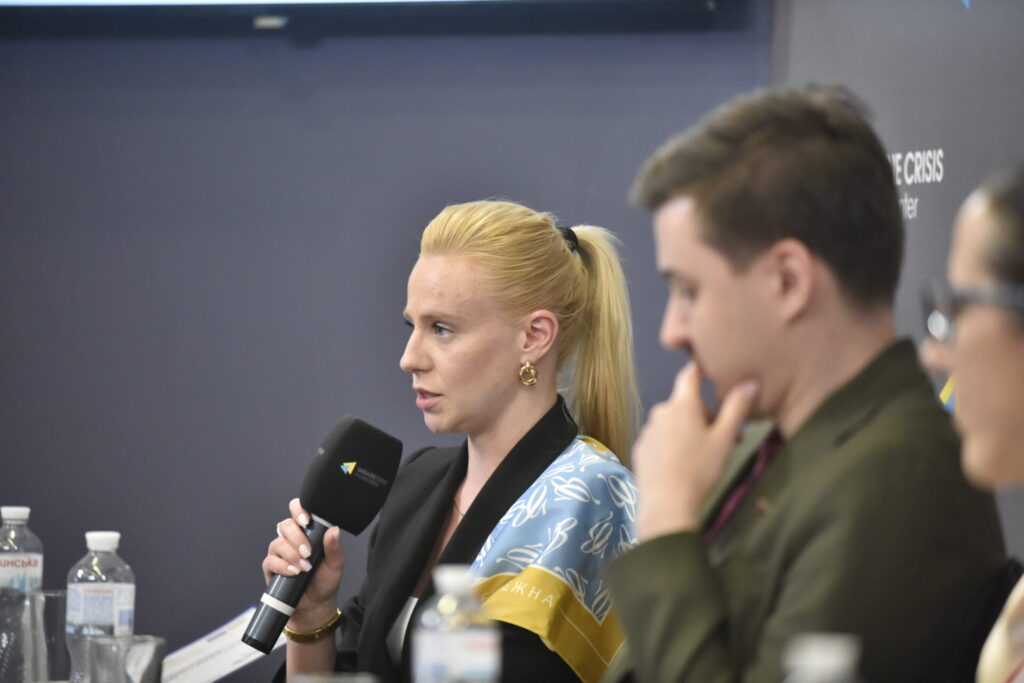 На фото в центрі у профіль Леся Хемраєва, керівниця відділу політики безбар'єрності, гендерної рівності та ментального здоров'я Міністерства культури та стратегічних комунікацій України . Вона - жінка зі світлим волоссям, зібраним у хвіст, у руках тримає мікрофон, на плечі костюму в неї жовто-блакитна шовкова хустина. Праворуч від неї, не у фокусі кадру, Вадим Міський і Юлія Сачук