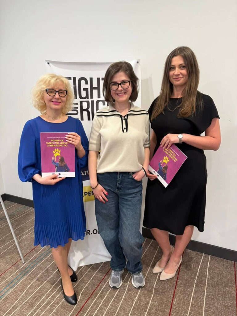 на фото три усміхнені жінки - Оксана Тулай, Єлизавета Борискіна і Євгенія Шелест. У Оксани, яка стоїть ліворуч, світле волосся до плечей, на ній синя класична сукня трохи нижче колін, у руках вона тримає посібник в темно-рожевій обкладинці "Розвиток лідерства дівчат з інвалідністю". У Євгенії, темноволосої жінки, яка стоїть праворуч у чорній класичній сукні, теж в руках такий самий посібник. Посередині на Єлизаветі світла блуза і сині джинси.
