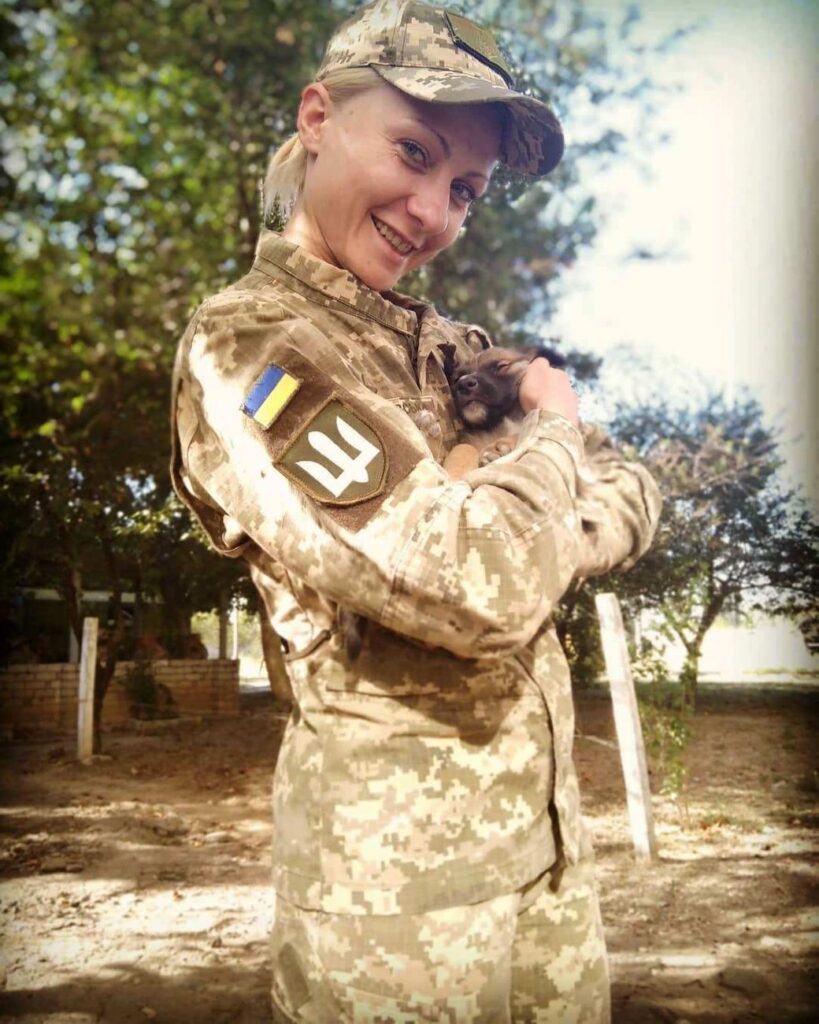 На фото молода усміхнена жінка зі світлим волоссям, зібраним у хвіст. Одягнена у військову форму з шевроном Сухопутних військ, що нагадує тризуб (білим кольором на темнозеленому фоні) на рукаві. Жінка тримає на руках цуценя. Це Анастасія Войнаровська, ветеранка, психологиня. 