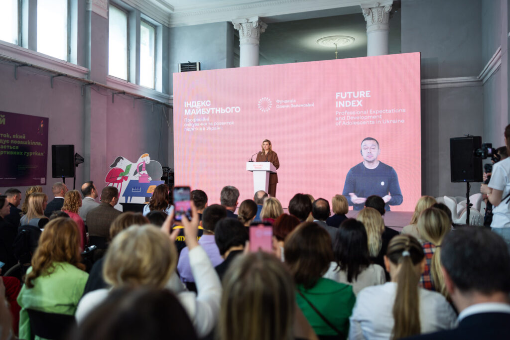 Олена Зеленська виступає під час презентації. Вона одягнена у коричневий костю. У неї світле коротке волосся. Позаду неї - банер рожевого кольору з назвою події. На банері відображається перекладач жестової мови. В залі сидять гості презентації. 