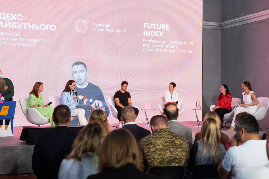 Юлія Сачук виступає під час презентації. Вона сидить на білому стільці-кріслі. Юлія одягнена у червону сорочку з білими смужками та довгими рукавами, чорні штани та туфлі. У неї довге темне волосся, вона носить окуляри. В руках вона тримає мікрофон. Поруч сидить у стільці-кріслі ведуча Катерина Осадча. Вона одягнена у білий костюм з коротким рукавом та у світлих туфлях. Її волосся заплетене в косу. В руках вона тримає мікрофон та дивиться на Юлію.