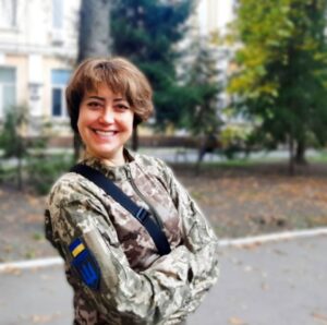 На фото усміхнена війська з коротким темно-русявим волоссям, у літній піксельній українській формі ЗСУ. На фоні видніється кількаповерхова споруда з блідо-жовтими стінами і дерева перед цією будівлею. 