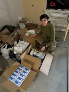 На фото усміхнена військова з темним волоссям, зібраним у хвіст, присіла біля картонних коробок з медикаментами. Біля неї з десяток коробок різних форм і величин, пакет. Жінка одягнена в камуфляжні штани і гольф зеленого кольору. На лівій руці в неї годинник чорного кольору 