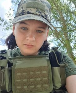 На селфі військова, на ній кепка піксельної літньої форми ЗСУ, темно-зелена футболка, поверх якої плитоноска з карманами, в яких видніються чорні джгути для зупинки кровотеч. На фоні дерева з зеленим листям і небо, вкрите хмарами.