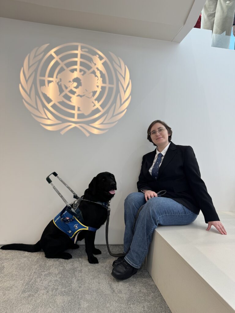 На фото Vikki чорний лабрадор супроводу і Катерина Щербина в темному костюмі, синій краватці поверх білої сорочки та джинсах сидить на парапеті біля знаку ООН на стіні.