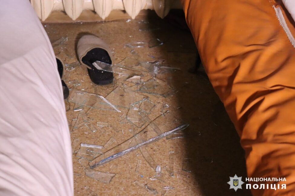 Фото Національної поліції в Харківській області. На фото коричневі капці під ламками скла стоять біля батареї, а обабіч них ліжка з постіллю блідорожевого (ліворуч) і теракотового (праворуч) кольорів