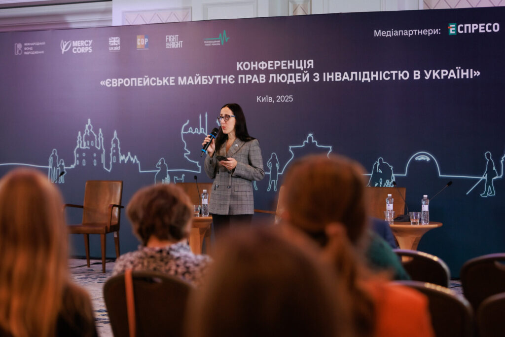 Юлія Сачук, голова Fight For Right, з вітальним словом перед учасниками та учасницями Конференції. Юлія в картатому чорно-білому жакеті з мікрофоном у руках на фоні банера Конференції "Європейське майбутнє прав людей з інвалідністю в Україні"