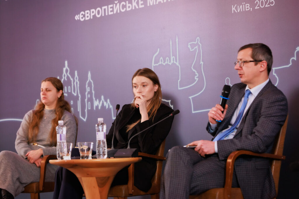 Людмила Фурсова (ліворуч) спікерка Конференції. Вона у сірій гарній сукні з тонким акцентним світлим поясом на талії. у зоні декольте золотистий ланцюжок з підвіскою. У Людмили довге русяве хвилясте волосся. Поруч (по центру) Вікторія Мельник, координаторка напряму «Європейська інтеграція» Центру політико-правових реформ, координаторка ЄвроХабу. Вона в темному жакеті, з довгим русявим волоссям прямим. Тримає руку біля губ, наче в задумі. Праворуч на фото Олександр Ільков, у сірому костюмі, світлій сорочці з блакитною краваткою. Він тримає мікрофон і говорить. Перед панелістами невисокий дерев'яний столик з двома склянками і двома пляшками води.