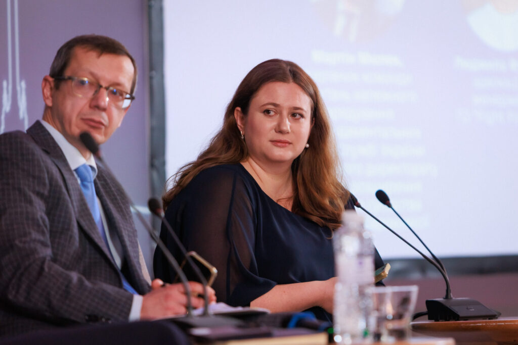 Ольга Лимар, виконавча директорка Коаліції РПР сидить праворуч у темній блузі, з довгим русявим волоссям. Перед нею тонкі два мікрофони. За нею видніється монітор. Поруч біля Ольги сидить Олександр Ільков, Генеральний директор Урядового офісу координації європейської та євроатлантичної інтеграції. Він одягнений у сірий картатий костюм, світлу сорочку з блакитною краваткою.