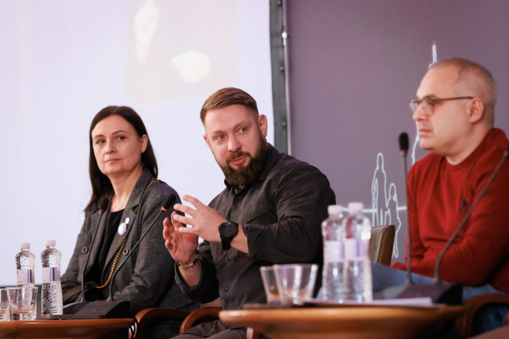 Зліва – направо: Тетяна Ломакіна, радниця-уповноважена Президента України з питань безбар'єрності. Одягнена у темну блузу, поверх якої сірий жакет. У Тетяни довге темне волосся. У центрі фото Руслан Приходько, заступник міністра у справах ветеранів України. Молодий чоловік зі стильною зачіскою, з вусами та бородою. Одягнений у темно-зелену сорочку. На лівій руці у нього чорний круглий годинник з чорним браслетом. Перед спікерами столики зі склянками та пляшками води.праворуч на фото (розмите зображення) Дмитро Луфер, консультант Міністерства охорони здоровʼя, експерт проєкту REHAB4U, одягнений у червоний светр. У нього коротке волосся з сивиною. Він носить окуляри з темними завушниками.