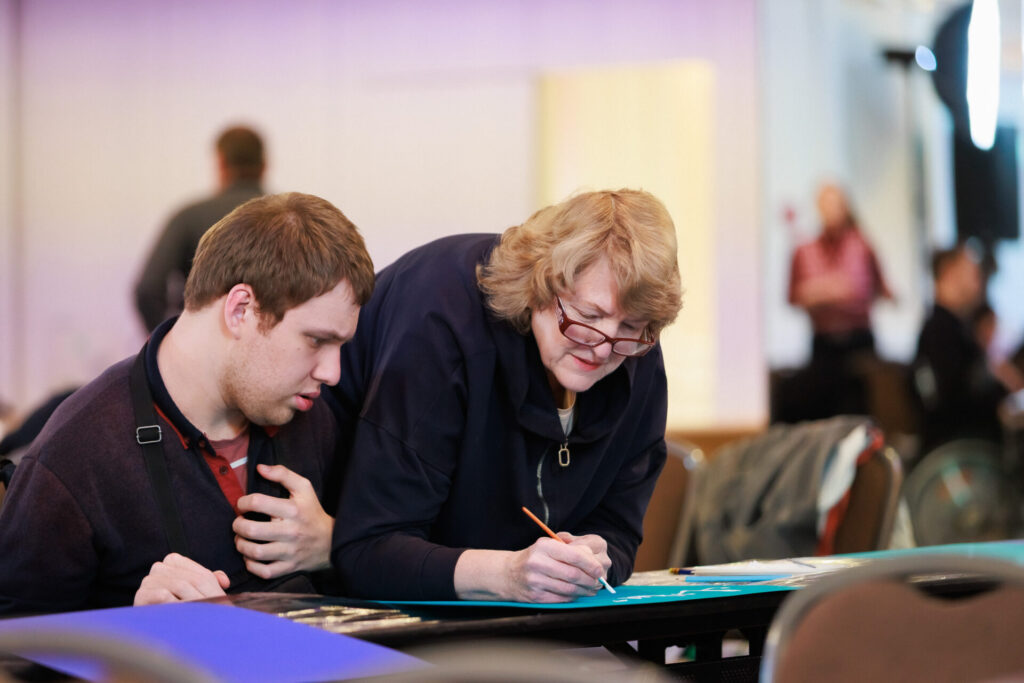 Учасники й учасниці готуються до Акції Lights for Rights. На фото зліва учасник Ігор в темному светрі, одягненому поверх червого реглану, молодий чоловік на кріслі колісному. Поруч із ним жінка старшого віку з коротким хвилястим світлим волоссям, в окулярах. Вона тримає в руках пензлик і пише ним по плакату темно-бірюзового кольору.