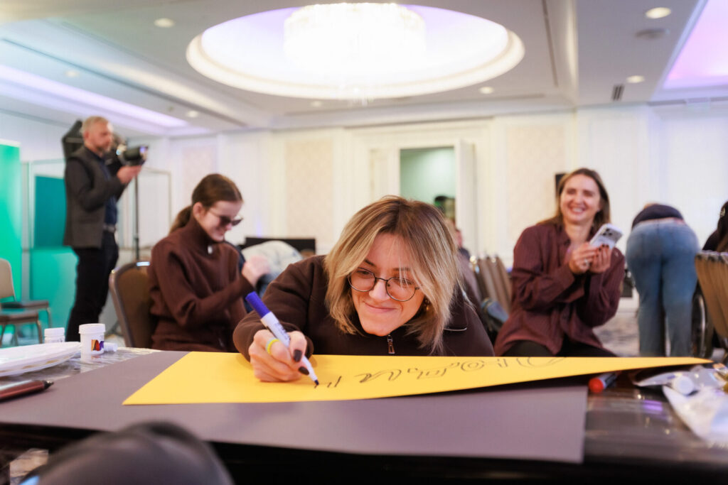 У центрі фото Алла Гінкул, молода усміхнена жінка зі світлим красивим волоссям, в окулярах. Вона готується до Акції Lights for Rights і пише свій лозунг на жовтому ватмані. За нею розмиті фігури двох інших учасниць - ліворуч Віра Семків, активістка, учасниця Школи "Лідерка-тінейджерка". Вона одягнена у коричневий костюм. А праворуч від Алли на фото Наталія Серватяк, депутатка й активістка зі Львова. Обидві мають довге волосся каштанового кольору. Наталія тримає в руках телефон світлого кольору. Скраю ліворуч за Вірою в русі потрапив у кадр перекладач жестової мови Ігор Шавро - високий чоловік з сивим волоссям, короткою бородою та вусами, одягнений у темні брюки та сірий жакет поверх темної сорочки. Ще на фото вгорі у центрі видно яскравий овал люстри. 