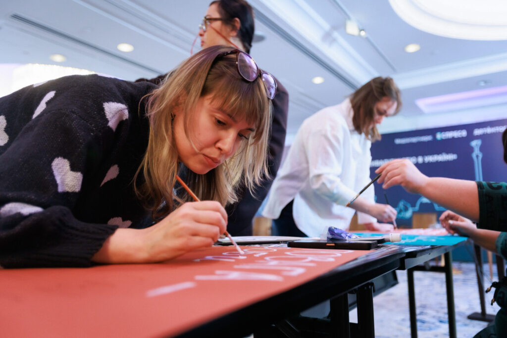 Валентина Пелюхня, активістка з Полтавщини малює власний плакат для Акції. У Валентини коротке світле волосся, зачіска з чубчиком. На голову жінка підняла окуляри. Одягнена у темний светр із білими сердечками.