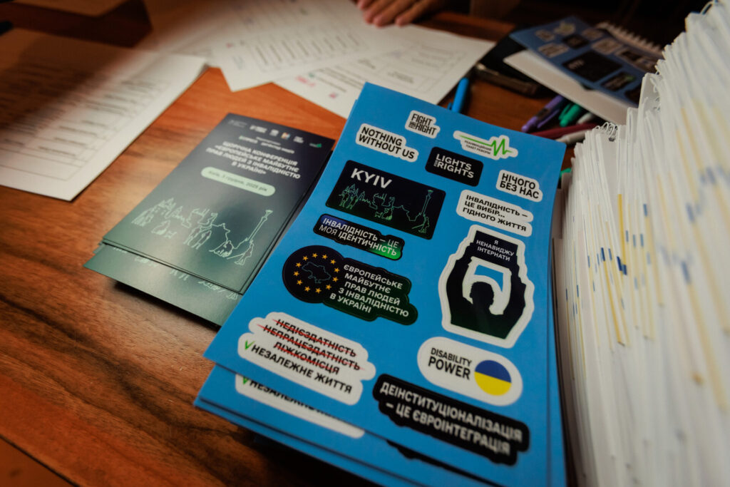 На столі, де відбувалася реєстрація учасників та учасниць Конференції, лежать наліпки з надписами про незалежне життя, видимість людей з інвалідністю тощо. А також флаєри з назвою Конференції та обрисами споруд Києва та обрисами людей з інвалідністю.